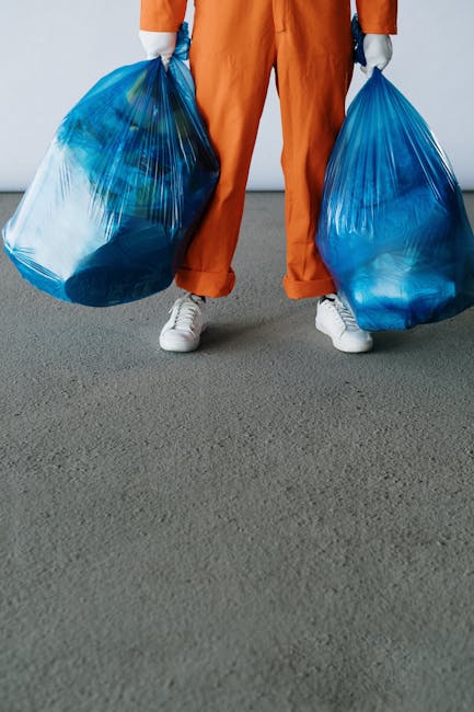 The image shows the lower half of a person standing on a gray carpeted floor, wearing orange work overalls and white sneakers. The individual is holding two large, semi-transparent blue plastic rubbish bags, one in each hand, filled with waste materials. The background is plain and neutral, likely an indoor environment such as a warehouse or storage area. The bags appear to contain various types of waste, possibly household or general rubbish, ready for disposal or collection. This scene suggests an independent waste removal activity, aligning with private rubbish collection services offered by companies like Waste Removal Croydon. The focus on the bags and the person's stance emphasizes the act of rubbish handling and on-site clearance, consistent with professional waste management or rubbish removal tasks undertaken outside of municipal collection practices.