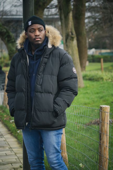 A young man with dark skin and a serious expression is standing outdoors next to a wire fence with wooden posts, in a green park or garden setting. He is wearing a black padded winter jacket with a faux fur trim around the hood, which is down, and a dark beanie hat. His hands are in the jacket pockets, and underneath, he has a navy blue hoodie. The background shows a grassy area with several large trees, some with bare branches, indicating a colder season. The environment is illuminated with natural daylight, giving a soft, diffuse light over the scene. The setting appears quiet and well-maintained, typical of a public park or garden area used for outdoor leisure activities, often the site for various waste and rubbish clearance services by companies like Waste Removal Croydon, which specialize in alternative waste handling and private disposal options away from local authority collections.