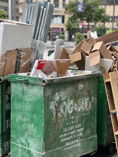 A close-up view of a large, weathered green waste disposal dumpster filled with various types of rubbish, including flattened cardboard boxes, some with printed graphics, white plastic containers, and miscellaneous packaging materials. The cardboard boxes are in shades of light brown, with some torn and partially collapsed, while others are neatly stacked or leaning against the side of the bin. The plastic containers are partially hidden among the boxes, visible near the top, with some crumpled and others intact. The background includes a blurred urban setting with trees, multi-storey buildings, and a few parked vehicles, indicating a city street or public area. The scene is illuminated by natural daylight, highlighting the textures of the worn, scratched, and slightly rusted metal surface of the dumpster, which has faded white writing and a partially obscured logo on its side. This image relates to private waste collection and rubbish removal services, showcasing the type of refuse managed during on-site clearance processes by companies such as Waste Removal Croydon.