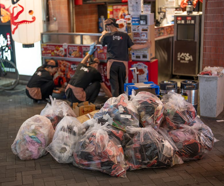 A collection of multiple large transparent plastic rubbish bags filled with mixed waste and textiles is situated on a paved sidewalk in front of a storefront, with some items visibly spilling out of the bags. Behind the bags, three workers, dressed in black uniforms with light brown aprons and gloves, are engaged in lifting or sorting waste. They are kneeling or crouching on the pavement near the entrance of a Japanese restaurant or food outlet, which features traditional style red paper lanterns and illuminated signage. To the right, there are two large black and silver litter bins or waste containers, and a blue plastic storage crate is also present on the ground. The scene appears to be an organized waste collection or on-site clearance operation, potentially arranged by Waste Removal Croydon for private waste handling, with the environment illuminated by ambient lighting, suggesting an evening setting. The surrounding area includes a brick façade and patio elements typical of urban commercial districts, emphasizing a professional approach to rubbish removal and disposal outside retail premises or event venues.