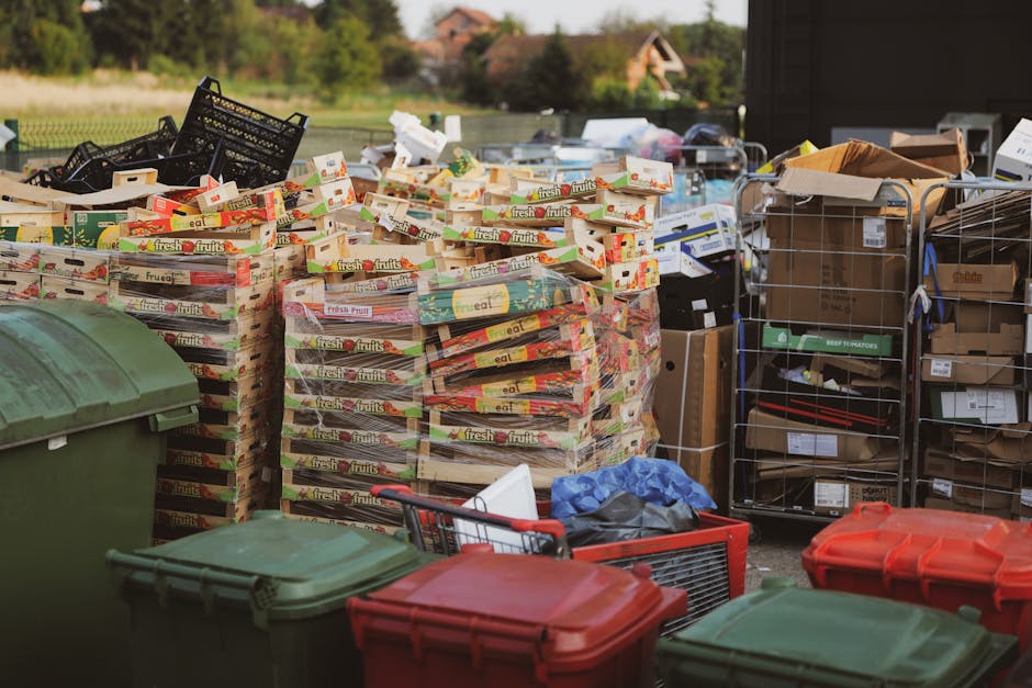 A close-up view of a large, weathered green waste disposal dumpster filled with various types of rubbish, including flattened cardboard boxes, some with printed graphics, white plastic containers, and miscellaneous packaging materials. The cardboard boxes are in shades of light brown, with some torn and partially collapsed, while others are neatly stacked or leaning against the side of the bin. The plastic containers are partially hidden among the boxes, visible near the top, with some crumpled and others intact. The background includes a blurred urban setting with trees, multi-storey buildings, and a few parked vehicles, indicating a city street or public area. The scene is illuminated by natural daylight, highlighting the textures of the worn, scratched, and slightly rusted metal surface of the dumpster, which has faded white writing and a partially obscured logo on its side. This image relates to private waste collection and rubbish removal services, showcasing the type of refuse managed during on-site clearance processes by companies such as Waste Removal Croydon.