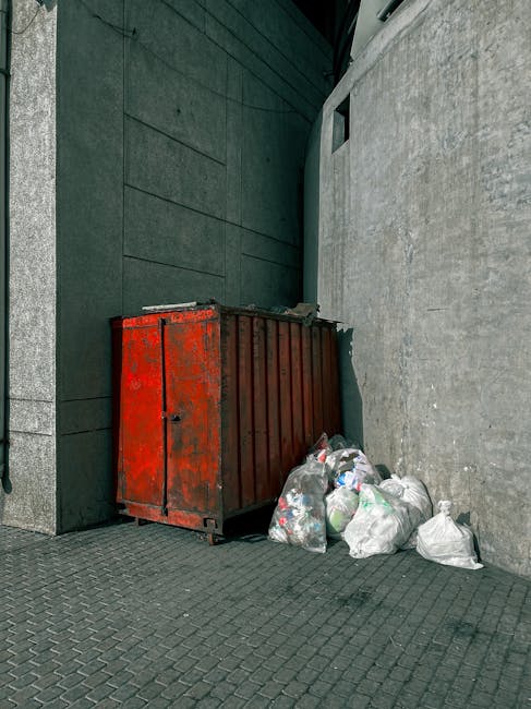 A large, weathered red metal dumpster positioned in a narrow corner between two building walls, one wall appearing dark green and textured, the other a light grey concrete surface. The dumpster is situated on a paved surface with a pattern of small, square, dark grey bricks. Several white plastic bags filled with rubbish are piled next to the dumpster, some leaning against it, with the bags partly transparent, revealing assorted waste inside. The scene is outdoors, likely on a street or alleyway, with minimal natural light casting soft shadows. The setting suggests an area for private waste disposal or bulk rubbish collection, reflecting typical rubbish removal practices supported by Waste Removal Croydon, a company specializing in rubbish services. The environment appears clean but shows signs of recent waste accumulation, highlighting the need for regular rubbish clearance in accordance with local council rules and regulations for bulky waste disposal.