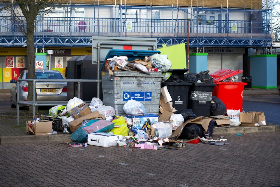 A large pile of mixed domestic waste and rubbish bags is accumulated outside on a paved urban sidewalk, surrounding multiple wheelie bins, including a grey bin labeled for mixed paper and card, a black bin for general waste, and a bright red recycling bin, with some bags and cardboard boxes spilling onto the ground. The rubbish appears to consist of discarded packaging, paper, cardboard, plastic items, and other household refuse, some items partially opened or crumpled. The scene is set against the backdrop of a building with a blue metal scaffolding structure covering its upper level and storefronts at street level. A silver car is parked nearby, partially obscured by metallic safety barriers. The lighting suggests daylight, contributing to a clear and detailed view of the overflowing rubbish, indicative of a collection issue or private waste disposal scenario managed by waste removal services, such as Waste Removal Croydon, in an urban environment.