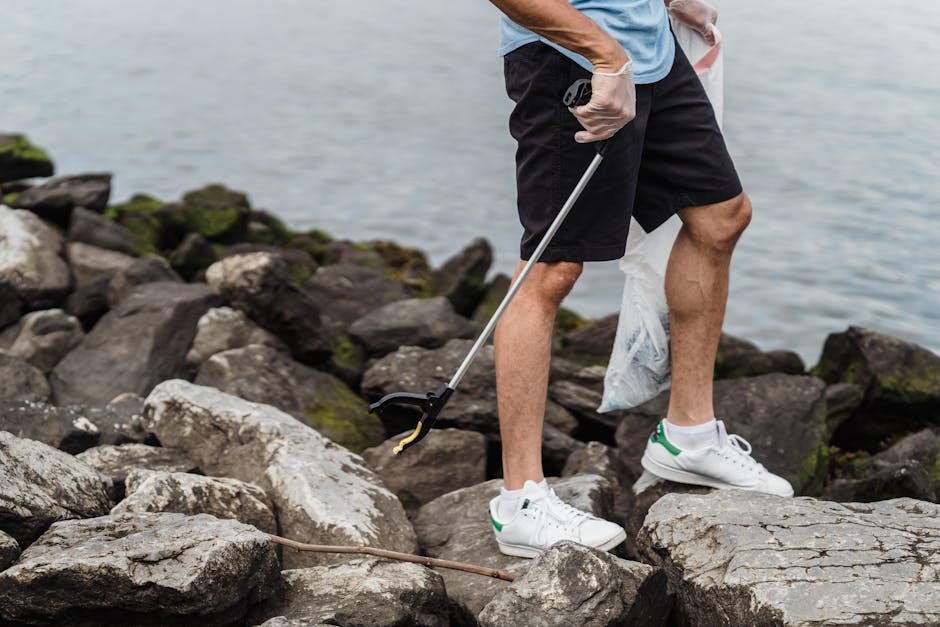 A person standing on a rocky shoreline near water, wearing casual clothing that includes a light blue t-shirt, black shorts, and white sneakers with green accents. They are holding a trash picker tool in their right hand, which is extended toward the ground, and a transparent plastic bag partially filled with litter in their left hand. The rocks around them are irregularly shaped with rough textures and various shades of grey and brown, some covered in patches of green moss. The water in the background appears calm, with a subtle sheen on its surface, and there are no other objects or people visible in the scene. The environment suggests an outdoor cleanup activity, which aligns with waste management and rubbish removal services like those offered by Waste Removal Croydon, often involving on-site collection and private disposal of debris and litter from natural or urban outdoor areas.