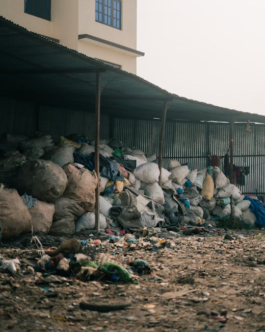 A large collection of mixed waste bags and loose rubbish is piled up beneath a corrugated metal shelter located outdoors adjacent to a building with beige walls and multiple windows. The waste bags are predominantly made of plastic, with some appearing transparent or semi-transparent, revealing various contents such as clothing, paper, and other household refuse. Several large, irregularly shaped sack-like bags are stacked along the length of the shelter, with some leaning or resting against a panelled fence that encloses the area. The ground in the foreground is uneven, covered with dirt, scattered debris, and smaller pieces of rubbish including plastic bottles, paper, and discarded items. The environment appears to be an informal or unmanaged waste storage area, which aligns with private rubbish collection or alternative waste handling services often provided by waste removal specialists like Waste Removal Croydon. The overall scene highlights the need for proper waste management solutions and demonstrates what might be seen during on-site clearance or private disposal efforts, with natural daylight illuminating the cluttered scene from an overcast sky.