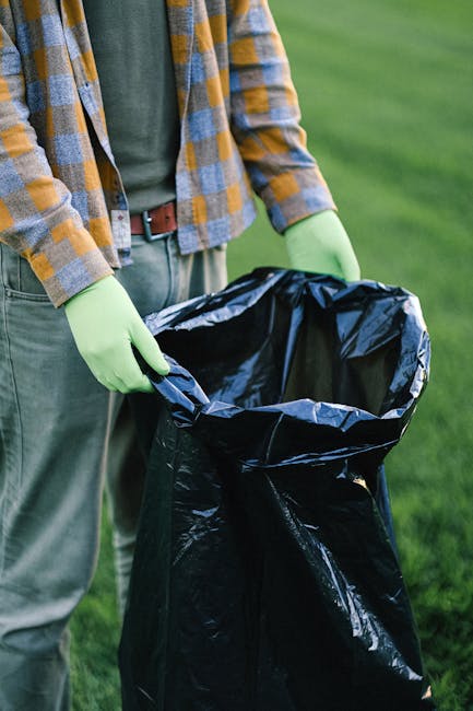 A person wearing a casual outfit consisting of a yellow, grey, and white checked shirt over a green T-shirt, with grey trousers secured by a brown belt, is holding open a large black rubbish bag lined with a metallic inner layer. The person is wearing light green gloves and standing outdoors on a grassy area, with a blurred background of green lawn. The scene appears to depict a waste collection or rubbish removal activity, possibly related to garden or outdoor rubbish clearance, reflecting the context of private waste handling or on-site rubbish disposal. The black bag is empty, ready to be filled with waste material, and the overall setting suggests an organized approach to rubbish removal, typical of professional waste collection services like Waste Removal Croydon.