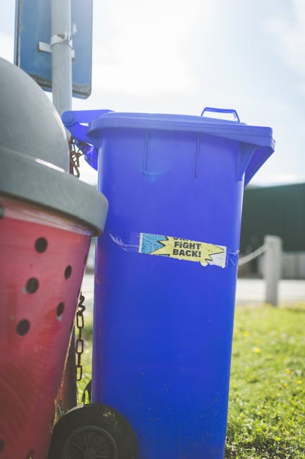 A tall, bright blue plastic rubbish bin with a closed lid, positioned on a grassy area outdoors. The bin is situated next to a pink bin with a black polka dot pattern and a gray lid, both partially visible in the foreground. The blue bin has a yellow and blue sticker with the text 'FIGHT BACK!' adhered to its front surface. In the background, there is a blue sign mounted on a pole, a grey metal bollard, and a road with guardrails under a partly cloudy sky, suggesting an outdoor setting potentially related to private waste collection or on-site rubbish disposal managed by waste removal services like Waste Removal Croydon. The lighting indicates daylight, with natural sunlight illuminating the scene and casting gentle shadows, emphasizing the textures of the plastic bins and the grass environment. This scene illustrates typical outdoor rubbish management, highlighting the use of dedicated bins for waste separation and collection.
