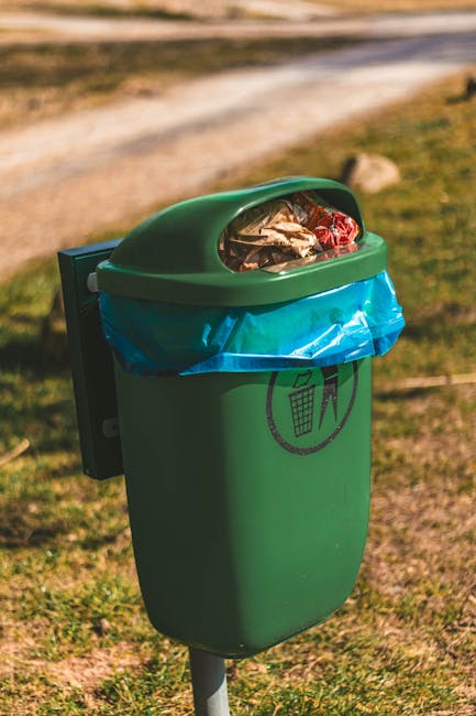 A person wearing a casual outfit consisting of a yellow, grey, and white checked shirt over a green T-shirt, with grey trousers secured by a brown belt, is holding open a large black rubbish bag lined with a metallic inner layer. The person is wearing light green gloves and standing outdoors on a grassy area, with a blurred background of green lawn. The scene appears to depict a waste collection or rubbish removal activity, possibly related to garden or outdoor rubbish clearance, reflecting the context of private waste handling or on-site rubbish disposal. The black bag is empty, ready to be filled with waste material, and the overall setting suggests an organized approach to rubbish removal, typical of professional waste collection services like Waste Removal Croydon.
